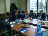 Grupa osób siedzi przy stole z dokumentami, poczęstunkiem oraz projektorem.