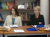 Dwie kobiety siedzą przy stole z niebieskimi teczkami oraz poczęstunkiem. W tle półki z książkami.