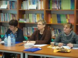 Trzy kobiety siedzą przy stole. W tle widać półki z książkami.