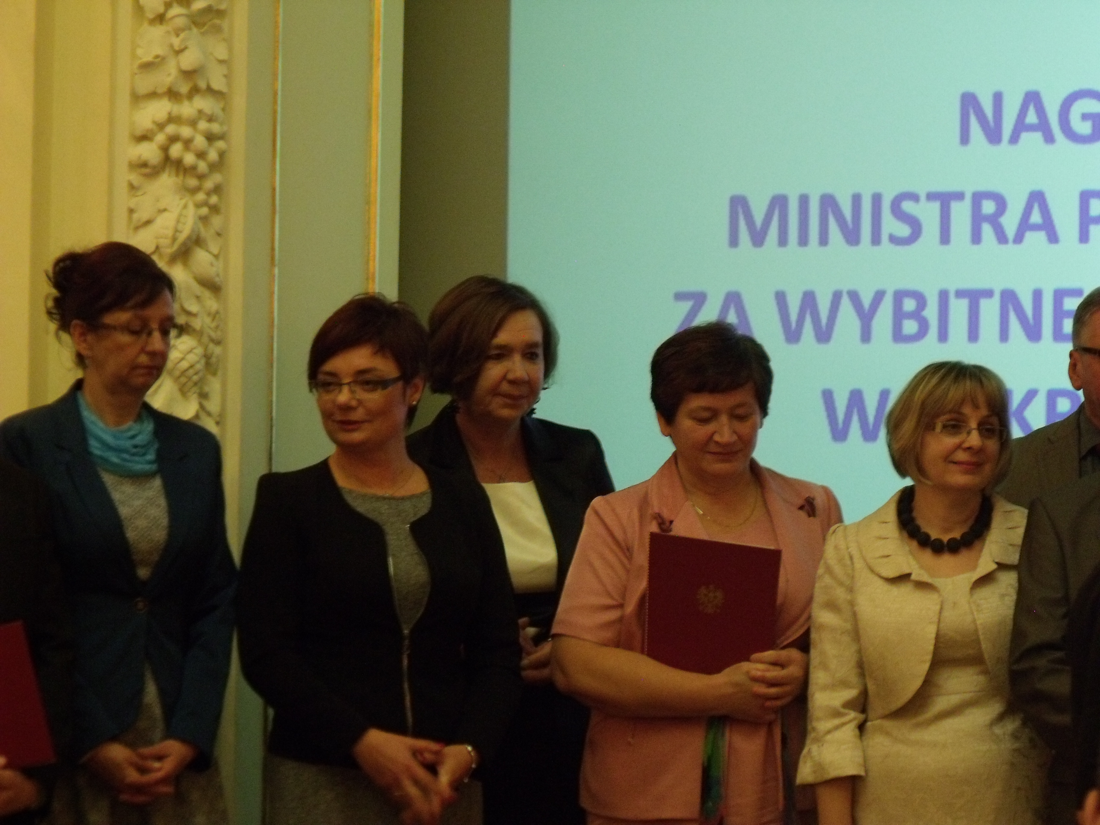 Grupa osób stoi obok siebie na tle prezentacji multimedialnej. Jedna z kobiet trzyma czerwoną teczkę.