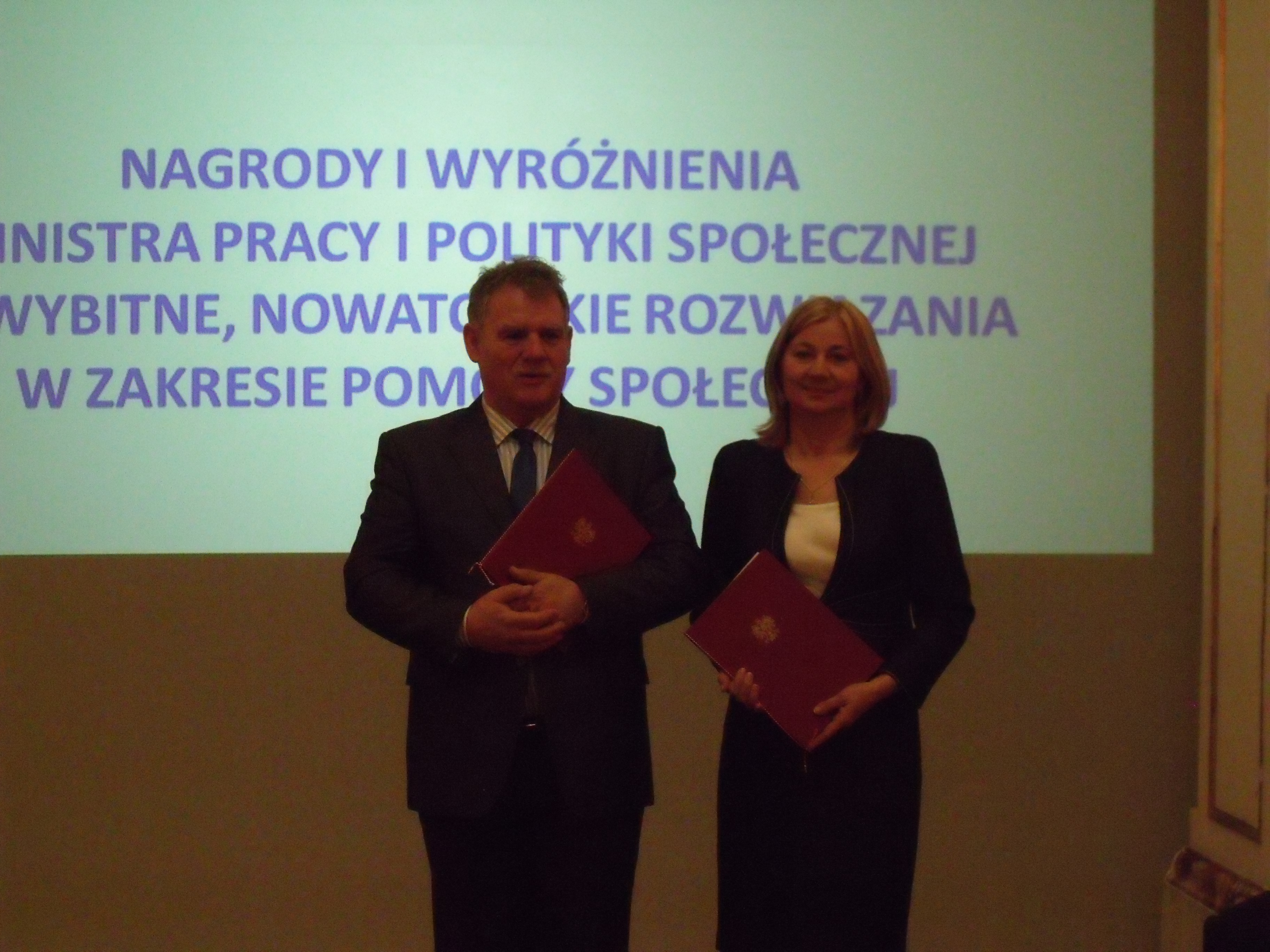 Dwie elegancko ubrane osoby z czerwonymi teczkami stoją na tle prezentacji multimedialnej.
