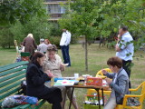 Grupa osób siedzi przy stołach pośród zieleni i maluje obrazy.