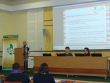 Trzy osoby prowadzące seminarium, z czego jedna przemawia na mównicy. W tle prezentacja multimedialna.