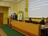 Grupa osób siedzi przy stole z ekranami tuż pod ekranem projekcyjnym. Obok przy mównicy stoi kobieta.