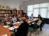 Grupa osób siedzi przy stole z dokumentami i poczęstunkiem. W tle widać półki z książkami i duże okno.
