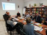 Grupa osób siedzi przy stole z dokumentami i poczęstunkiem. Na ścianie jest wyświetlony tekst z projektora.