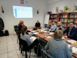 Grupa osób siedzi przy stole z dokumentami i poczęstunkiem. Na ścianie jest wyświetlony tekst z projektora.