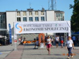 Duży plac na którym spacerują ludzie. Na środku placu stoi duży baner Podlaskich Targów Ekonomii Społecznej. Wokół stoją stoiska.
