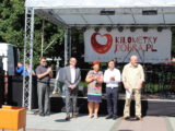 Pięć osób stoi w jednym rzędzie przy scenie i bije brawo. W tle widać sprzęt muzyczny oraz baner Kilometry Dobra.