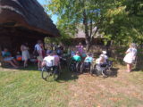 Grupa dorosłych z dziećmi na wózkach inwalidzkich zgromadzona pod drzewem przy płocie. Część osób siedzi pod dachem ze strzechy.