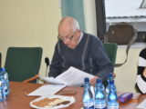 Starszy mężczyzna w okularach siedzi przy stole z poczęstunkiem i czyta dokument.