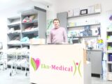Uśmiechnięty mężczyzna stoi za blatem wewnątrz placówki Eko-Medical która zajmuje się sprzętem medycznym. Wokół na półkach leżą różne artykuły medyczne.