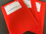 Czerwone teczki z gumką z emblematami funduszów europejskich i zatytułowane Projekt partnerski Akademia Pomocy Społecznej.