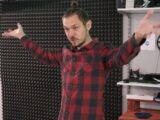 Mężczyzna z uniesionymi rękoma stoi na tle ściany z pianką akustyczną i innym sprzętem typ telewizor i głośniki.