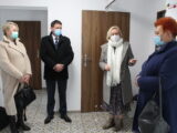 Cztery osoby w maseczkach stoją przy ścianach w jasnym pomieszczeniu z drzwiami. Jedna z kobiet gestykuluje.