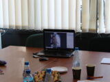Laptop stoi na końcu stołu na którym znajdują się dokumenty i poczęstunek. Na laptopie jest wyświetlony mężczyzna ze spotkania online.