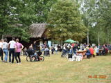 Piknik integracyjny dla osób niepełnosprawnych i ich rodzin. Tłum ludzi zebrany na dużym, zielonym podwórku z domem krytym strzechą. Kilka osób siedzi przy stolikach.
