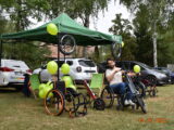 Piknik integracyjny dla osób niepełnosprawnych i ich rodzin. Mężczyzna przed zielonym namiotem siedzi na wózku inwalidzkim z przystawką elektryczną. Obok stoją jeszcze dwa takie wózki. W tle stoją samochody.