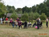 Piknik integracyjny dla osób niepełnosprawnych i ich rodzin. Grupa opiekunów, dzieci oraz osób na wózkach inwalidzkich na zielonej trawie. W dalszym planie stoi kilka osób przy stołach. W tle widać dom kryty strzechą.