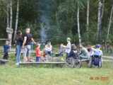 Piknik integracyjny dla osób niepełnosprawnych i ich rodzin. Grupa dorosłych, dzieci i osób na wózkach inwalidzkich pieczę kiełbaski nad ogniskiem. W tle stoi rower, a w dalszym planie rośnie gęsty, zielony las.