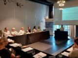 Sala z stołami ustawionymi w kształt litery U przy których siedzą uczestnicy. Na środku jest wyświetlona prezentacja. Pod ekranem projekcyjnym stoi stół przy którym siedzi mężczyzna prowadzący spotkanie.