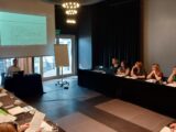 Sala z stołami ustawionymi w kształt litery U przy których siedzą uczestnicy. Na środku jest wyświetlona prezentacja. Pod ekranem projekcyjnym stoi stół przy którym siedzi mężczyzna prowadzący spotkanie.