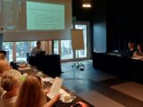 Sala z wyświetloną prezentacją na środku sali. Pod ekranem projekcyjnym stoi stół przy którym siedzi mężczyzna prowadzący spotkanie. Z lewej i prawej strony przy podłużnych stołach siedzą uczestnicy spotkania.