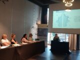 Sala z wyświetloną prezentacją na środku sali. Pod ekranem projekcyjnym stoi stół przy którym siedzi kobieta prowadząca spotkanie. Z lewej strony przy podłużnym stole siedzą uczestnicy spotkania.