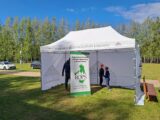 Namiot Podlaskie Muzeum Kultury Ludowej. W środku stoi dorosły, dwójka dzieci oraz baner ROPS w Białymstoku. Wokół dużo zieleni.