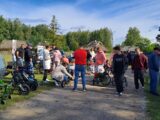 Zgromadzenie ludzi na piaszczystej drodze. Wśród nich są dzieci na wózkach inwalidzkich. W tle dużo zieleni, drzew i kilka drewnianych domów.