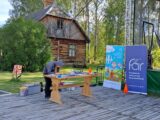 Drewniany podest na którym stoi drewniany stół przy którym pochyla się kobieta. Obok stoją 2 banery. Obok stoi scena a w tle stoi drewniany dom.