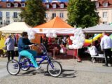 Stoiska podmiotów ekonomii społecznej wystawione w jednym rzędzie na rynku Kościuszki w Białymstoku z okazji Targów Ekonomii Społecznej. Przed stoiskami przejeżdża mężczyzna na rowerze trójkołowym.