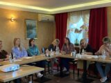 Sala z łączonymi stołami w kształt litery L przy których siedzą uczestnicy szkolenia. Na środku stoi mały stolik z projektorem.
