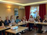 Sala z łączonymi stołami w kształt litery L przy których siedzą uczestnicy szkolenia. Na środku stoi mały stolik z projektorem.