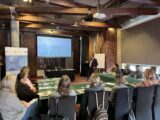 Grupa osób siedzi przy łączonych stołach w trakcie szkolenia. Przed nimi stoi prowadzący, który pokazuje coś na ekranie projekcyjnym.