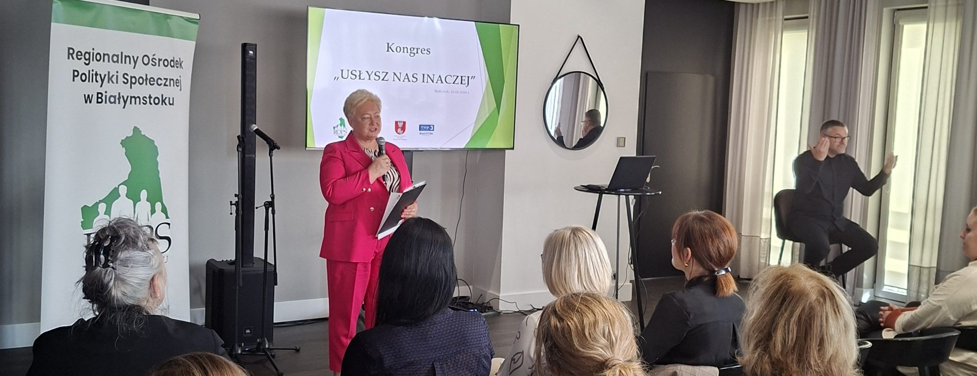 Dyrektor Regionalnego Ośrodka Polityki Społecznej w Białymstoku Pani Bożena Tomaszewska przemawia przed publicznością. Za nią wyświetlona prezentacja na ekranie oraz baner ROPS. Obok siedzi mężczyzna który tłumaczy na migowy. Miniatura zdjęcia.